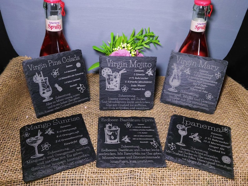 May include: Six black slate coasters with engraved cocktail recipes, including Virgin Pi&ntilde;a Colada, Virgin Mojito, Virgin Mary, Mango Sunrise, Erdbeer Basilikum-Spritz, and Ipanema. The coasters are arranged on a burlap surface, with two bottles of red liquid in the background.