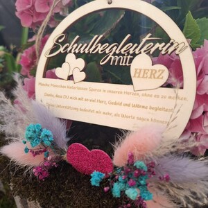 Op de afbeelding: Een houten, cirkelvormige plaque met de woorden "Schulbegleiterin mit HERZ" uitgesneden. De plaque is versierd met twee hartvormen en een glinsterend roze hart. De plaque is omgeven door roze bloemen en kleurrijke gedroogde bloemen.