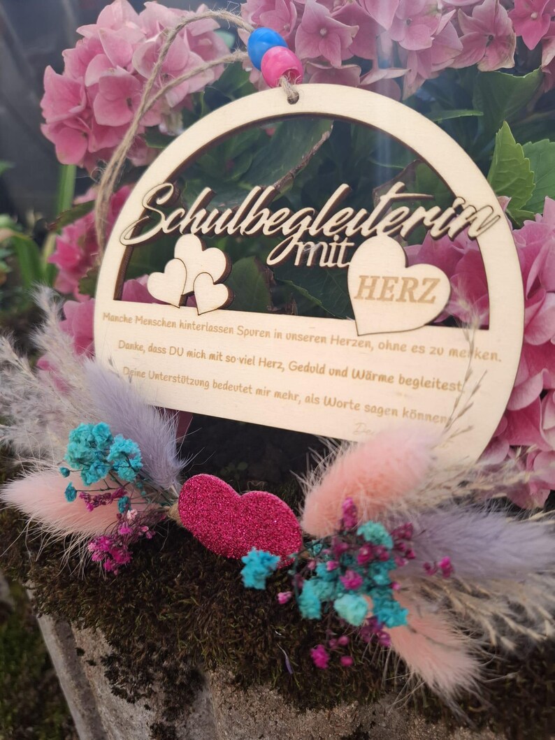 Op de afbeelding: Een houten, cirkelvormige plaque met de woorden "Schulbegleiterin mit HERZ" uitgesneden. De plaque is versierd met twee hartvormen en een glinsterend roze hart. De plaque is omgeven door roze bloemen en kleurrijke gedroogde bloemen.