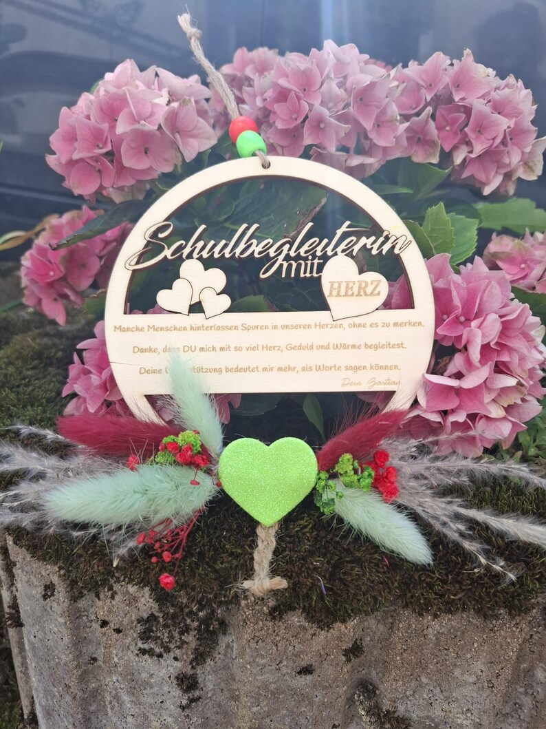 Op de afbeelding: Een houten cirkelvormig ornament met de woorden "Schulbegleiterin mit HERZ" en een oprechte boodschap. Het ornament heeft hartvormige uitsparingen en is versierd met een groen hart, rode en groene accenten en roze bloemen.
