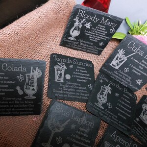 May include: A collection of black slate coasters with engraved cocktail recipes, including Pina Colada, Bloody Mary, and Tequila Sunrise. The coasters feature detailed illustrations of the drinks and their ingredients, with white text on a dark background.