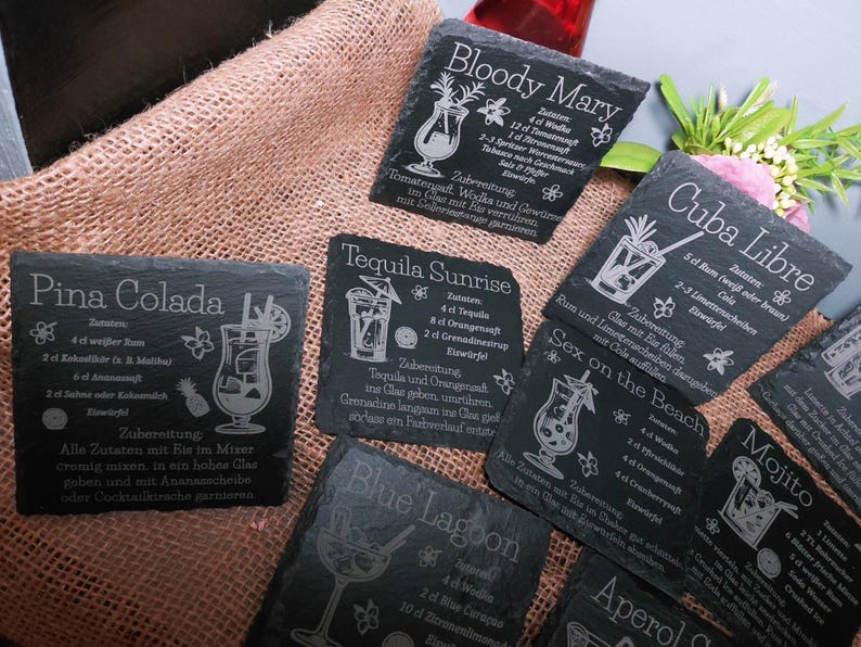 May include: A collection of black slate coasters with engraved cocktail recipes, including Pina Colada, Bloody Mary, and Tequila Sunrise. The coasters feature detailed illustrations of the drinks and their ingredients, with white text on a dark background.