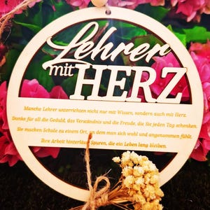 Op de afbeelding: Een houten cirkelvormig ornament met de Duitse tekst "Lehrer mit HERZ" (Leraar met HART). Het ornament is omgeven door roze bloemen en heeft een klein boeket gedroogde bloemen vastgebonden met touw.