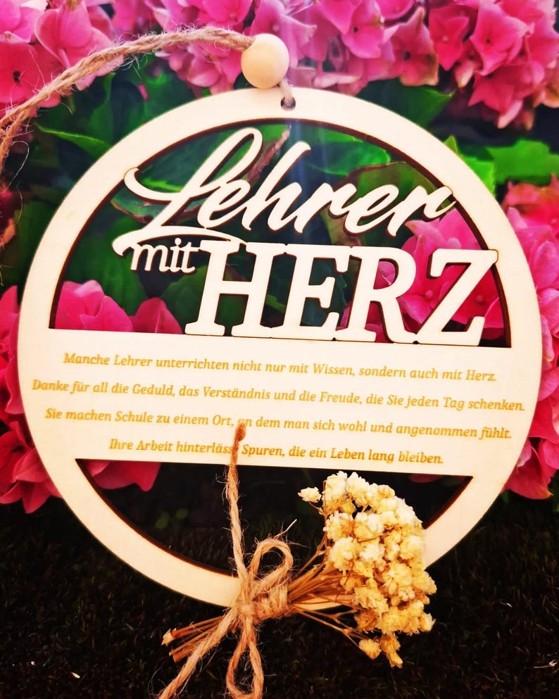 Op de afbeelding: Een houten cirkelvormig ornament met de Duitse tekst "Lehrer mit HERZ" (Leraar met HART). Het ornament is omgeven door roze bloemen en heeft een klein boeket gedroogde bloemen vastgebonden met touw.