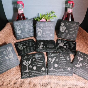 May include: A collection of slate coasters engraved with cocktail recipes, including "Bloody Mary," "Cuba Libre," and "Caipirinha." The coasters are arranged on a burlap surface, with two bottles of red soda in the background.