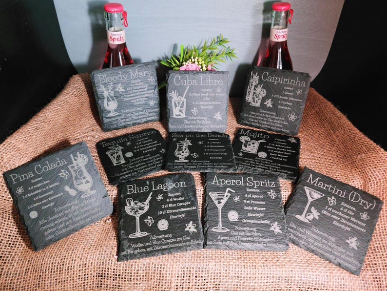 May include: A collection of slate coasters engraved with cocktail recipes, including "Bloody Mary," "Cuba Libre," and "Caipirinha." The coasters are arranged on a burlap surface, with two bottles of red soda in the background.