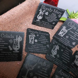 May include: A collection of black slate coasters with engraved cocktail recipes, including Pina Colada, Bloody Mary, and Tequila Sunrise. The coasters feature detailed illustrations of the drinks and their ingredients, with text in white.