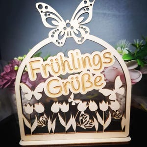 May include: Wooden decorative piece with a butterfly on top and the words "Frühlings Grüße" in the center. The design includes butterflies and tulips. The piece is made of light-colored wood.