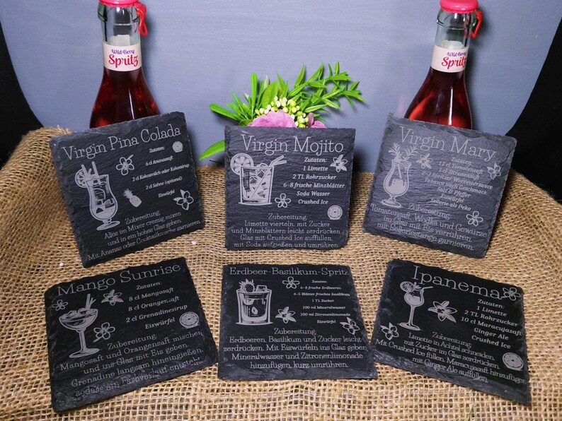 May include: Six black slate coasters with engraved cocktail recipes, including Virgin Pi&ntilde;a Colada, Virgin Mojito, Virgin Mary, Mango Sunrise, Erdbeer Basilikum-Spritz, and Ipanema. The coasters are arranged on a burlap surface, with two bottles of red liquid in the background.
