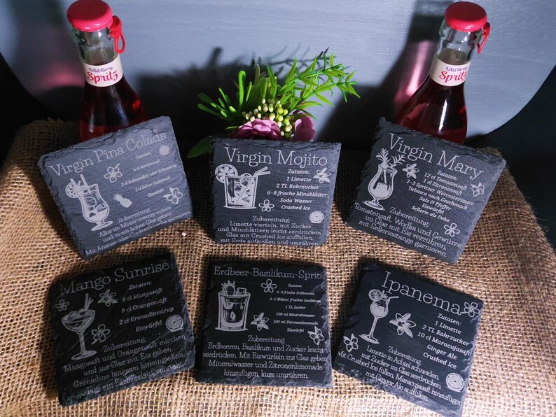 May include: A collection of six slate coasters with cocktail recipes engraved in white. The coasters include recipes for Virgin Pina Colada, Virgin Mojito, Virgin Mary, Mango Sunrise, Erdbeer-Basilikum-Sprit, and Ipanema. Two bottles of Spritz are in the background.