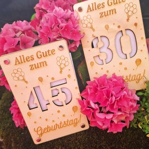 May include: Two wooden birthday signs with the text "Alles Gute zum Geburtstag!" and balloon illustrations. One sign displays the number 30, the other displays 45. The signs are surrounded by vibrant pink flowers.