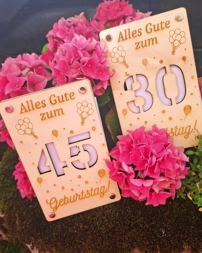 May include: Two wooden birthday signs with the text "Alles Gute zum Geburtstag!" and balloon illustrations. One sign displays the number 30, the other displays 45. The signs are surrounded by vibrant pink flowers.