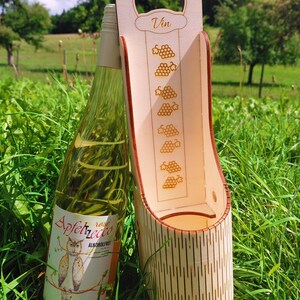 May include: A wooden wine bottle holder with a handle, featuring the word "Vin" and grape illustrations. A bottle of Apfelzecto wine with an owl label rests beside it, all set in green grass.