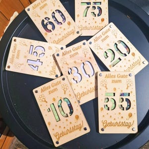 May include: Wooden birthday cards with laser-cut numbers 10, 20, 30, 45, 55, 60, and 75. Each card has the German text "Alles Gute zum Geburtstag!" and balloon graphics. The cards are made of light-colored wood.