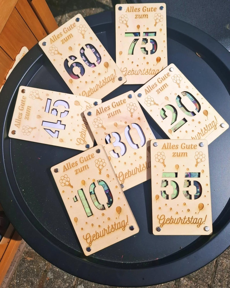 May include: Wooden birthday cards with laser-cut numbers 10, 20, 30, 45, 55, 60, and 75. Each card has the German text "Alles Gute zum Geburtstag!" and balloon graphics. The cards are made of light-colored wood.