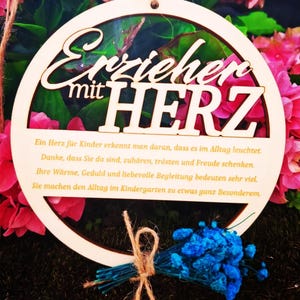 Op de afbeelding: Een houten, cirkelvormig bord met de Duitse tekst "Erzieher mit HERZ" (Opvoeder met hart). Het bord is omgeven door roze bloemen en een klein boeket blauwe bloemen vastgebonden met touw.