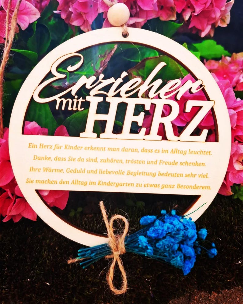 Op de afbeelding: Een houten, cirkelvormig bord met de Duitse tekst "Erzieher mit HERZ" (Opvoeder met hart). Het bord is omgeven door roze bloemen en een klein boeket blauwe bloemen vastgebonden met touw.