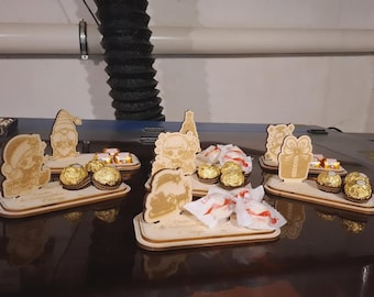 May include: Wooden laser-cut decorative trays with various designs, including gnomes, a girl, a Christmas tree, and a gift box. Each tray holds wrapped chocolates. The items are arranged on a dark surface.