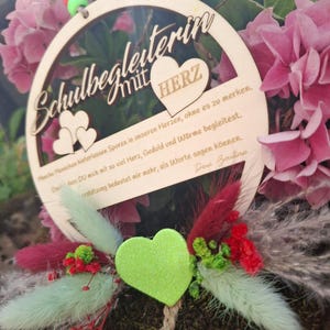 Op de afbeelding: Een houten cirkelvormig ornament met de Duitse tekst "Schulbegleiter mit HERZ" en een hartvormig ontwerp. Het ornament is omgeven door roze bloemen en decoratief groen, waaronder een groen hart en rode accenten.