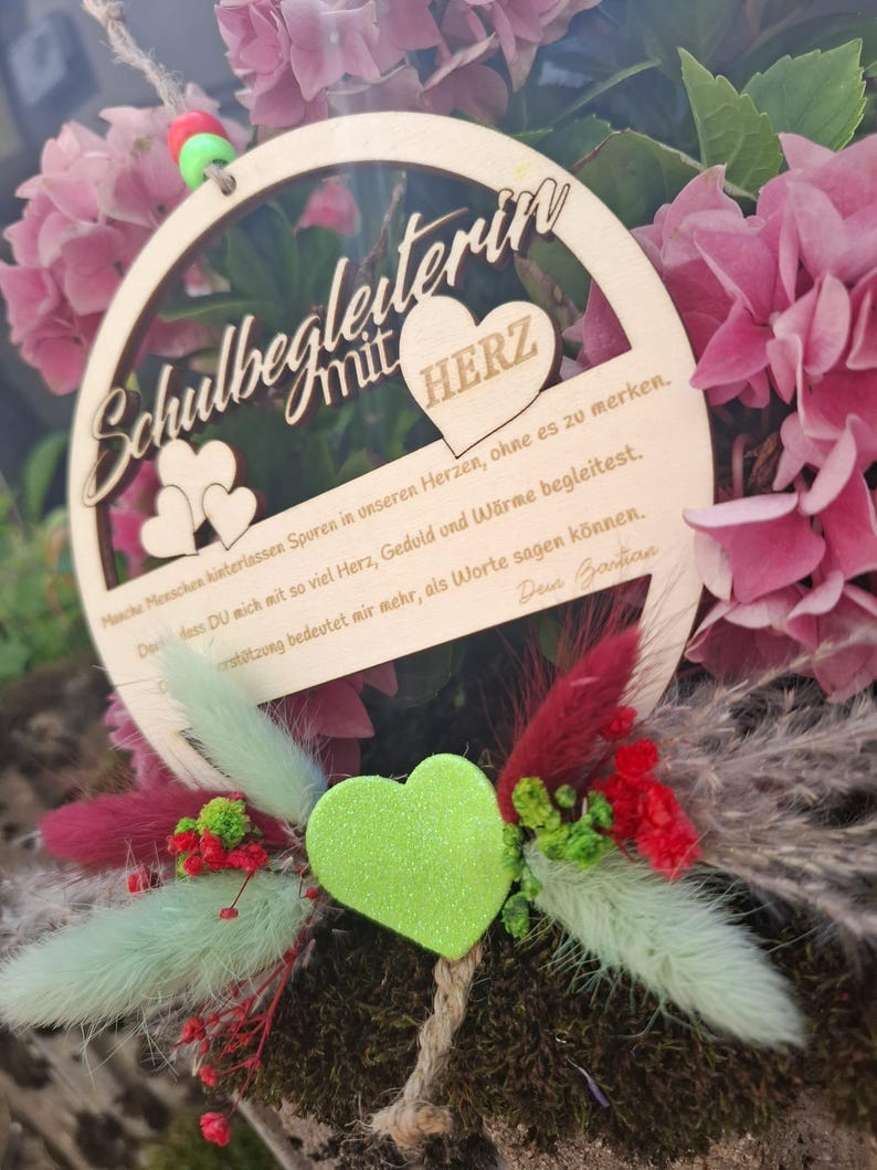 Op de afbeelding: Een houten cirkelvormig ornament met de Duitse tekst "Schulbegleiter mit HERZ" en een hartvormig ontwerp. Het ornament is omgeven door roze bloemen en decoratief groen, waaronder een groen hart en rode accenten.
