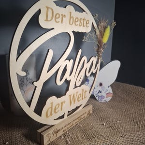 May include: A wooden sign with the words "Der beste Papa der Welt" (The best Dad in the world) cut out. The sign is round with a decorative floral element and sits on a wooden base. The base has additional text.