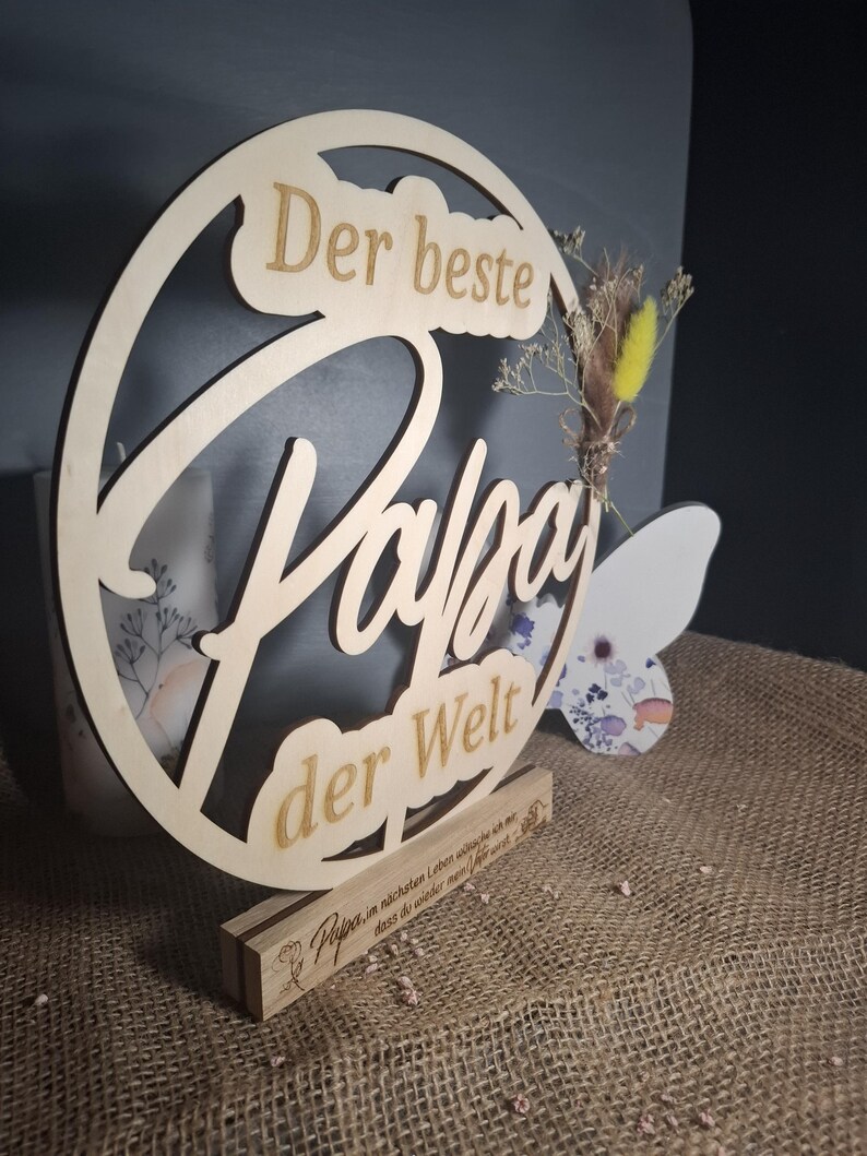 May include: A wooden sign with the words "Der beste Papa der Welt" (The best Dad in the world) cut out. The sign is round with a decorative floral element and sits on a wooden base. The base has additional text.