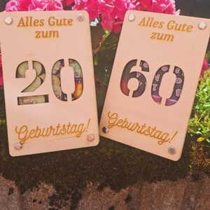 May include: Two wooden birthday cards with the text "Alles Gute zum Geburtstag!" The cards have cut-out numbers 20 and 60, with decorative elements visible through the cutouts. The cards are light brown and have small, round, silver-colored rivets.