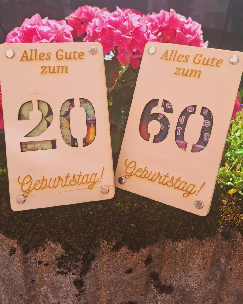 May include: Two wooden birthday cards with the text "Alles Gute zum Geburtstag!" The cards have cut-out numbers 20 and 60, with decorative elements visible through the cutouts. The cards are light brown and have small, round, silver-colored rivets.