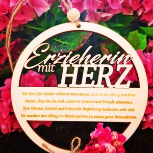 Op de afbeelding: Een houten, cirkelvormig ornament met de Duitse tekst "Erzieherin mit HERZ" (Educator met hart). Het ornament heeft een natuurlijke houten afwerking en wordt opgehangen door een touwlus. De achtergrond toont roze bloemen.