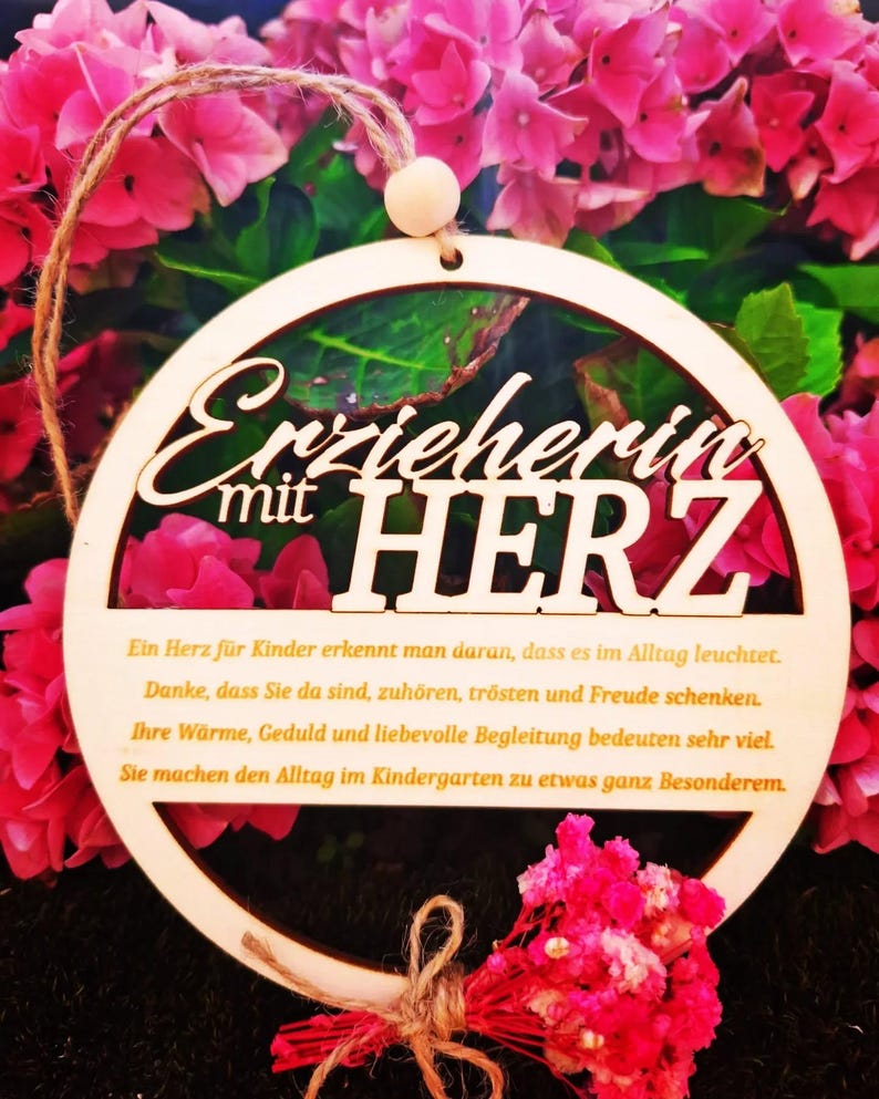 Op de afbeelding: Een houten, cirkelvormig ornament met de Duitse tekst "Erzieherin mit HERZ" (Educator met hart). Het ornament heeft een natuurlijke houten afwerking en wordt opgehangen door een touwlus. De achtergrond toont roze bloemen.
