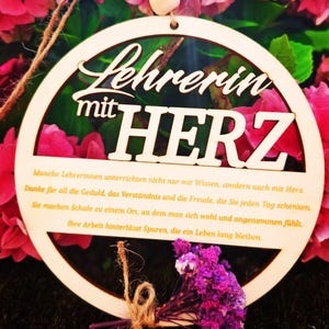 Op de afbeelding: Houten cirkelvormige plaque met de woorden "Lehrerin mit HERZ" uitgesneden. De plaque heeft een klein boeket paarse bloemen vastgebonden met touw. De achtergrond is een mix van roze bloemen en groene bladeren.