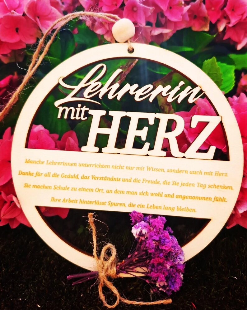 Op de afbeelding: Houten cirkelvormige plaque met de woorden "Lehrerin mit HERZ" uitgesneden. De plaque heeft een klein boeket paarse bloemen vastgebonden met touw. De achtergrond is een mix van roze bloemen en groene bladeren.
