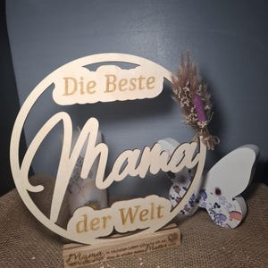 May include: A wooden sign with the words "Die Beste Mama der Welt" in a decorative font. The sign includes a small wooden base with text and a butterfly decoration. A small bundle of dried flowers is attached to the sign.
