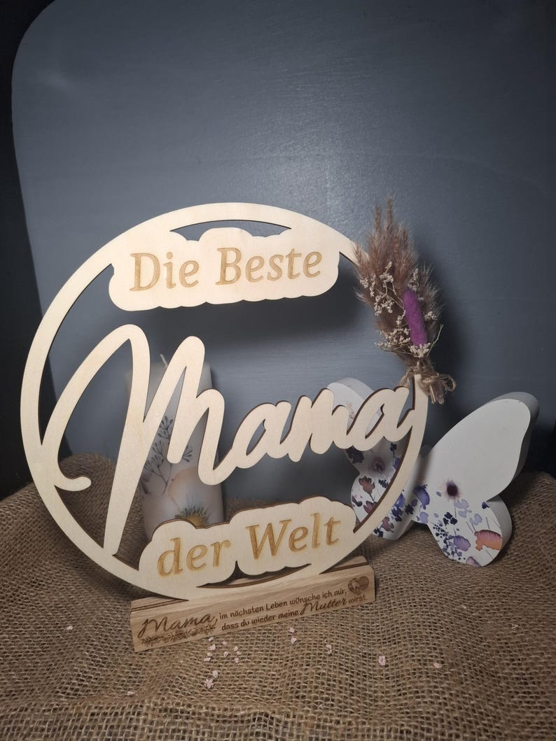 May include: A wooden sign with the words "Die Beste Mama der Welt" in a decorative font. The sign includes a small wooden base with text and a butterfly decoration. A small bundle of dried flowers is attached to the sign.