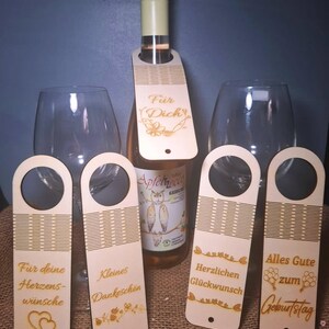 May include: A bottle of rosé wine with a wooden tag that says "Für Dich". Four additional wooden tags with various German phrases, including "Für deine Herzenswünsche" and "Alles Gute zum Geburtstag". Two wine glasses are visible.