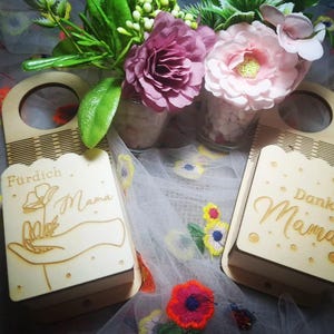 May include: Two wooden door hangers with a rectangular design and a circular handle. One says "Fürdich Mama" and the other says "Danke Mama" in cursive. The hangers are light brown with a floral design and small dots. Flowers are in the background.