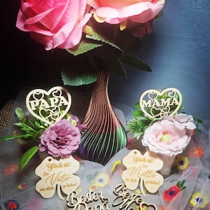 May include: A display of pink and yellow roses in a decorative vase. Wooden cutouts with the words "PAPA" and "MAMA" are visible, alongside clover-shaped tags with German text. The arrangement is set on a white fabric with floral patterns.
