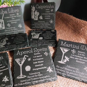 May include: A collection of black slate coasters with engraved cocktail recipes. The coasters feature recipes for drinks like Cuba Libre, Caipirinha, Mojito, Aperol Spritz, Lagoon, and Martini (Dry). Each coaster has a unique design.
