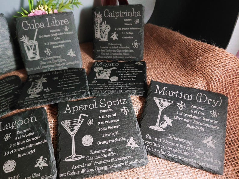 May include: A collection of black slate coasters with engraved cocktail recipes. The coasters feature recipes for drinks like Cuba Libre, Caipirinha, Mojito, Aperol Spritz, Lagoon, and Martini (Dry). Each coaster has a unique design.