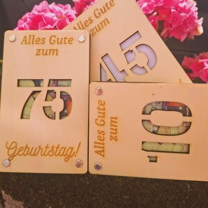 May include: Wooden birthday signs with cut-out numbers 10, 45, and 75, and the German phrase "Alles Gute zum Geburtstag!" in gold lettering. The signs are light brown with silver rivets and are displayed in front of pink flowers.