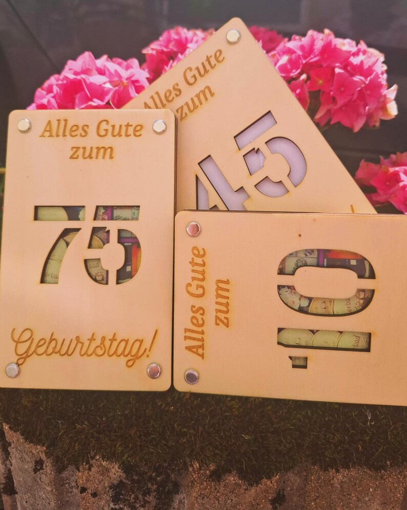 May include: Wooden birthday signs with cut-out numbers 10, 45, and 75, and the German phrase "Alles Gute zum Geburtstag!" in gold lettering. The signs are light brown with silver rivets and are displayed in front of pink flowers.