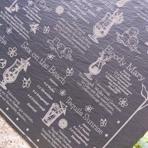 May include: A black slate serving board etched with cocktail recipes, including Martini, Pina Colada, Sex on the Beach, Bloody Mary, and Tequila Sunrise. Decorative floral and cocktail glass illustrations accompany the text.