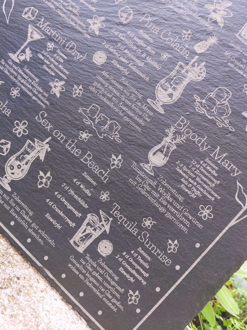 May include: A black slate serving board etched with cocktail recipes, including Martini, Pina Colada, Sex on the Beach, Bloody Mary, and Tequila Sunrise. Decorative floral and cocktail glass illustrations accompany the text.