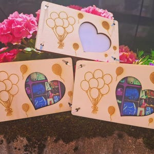 May include: Three wooden picture frames with a heart-shaped cutout and balloon designs. The frames are light-colored wood, with a laser-cut design of balloons and a heart shape. The background includes pink flowers and greenery.