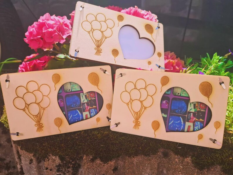 May include: Three wooden picture frames with a heart-shaped cutout and balloon designs. The frames are light-colored wood, with a laser-cut design of balloons and a heart shape. The background includes pink flowers and greenery.