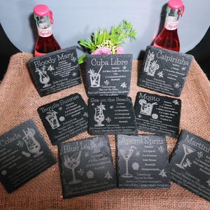May include: A collection of dark gray slate coasters with engraved cocktail recipes, including "Bloody Mary," "Cuba Libre," and "Mojito." The coasters are arranged on a brown burlap surface, with two bottles of pink soda in the background.