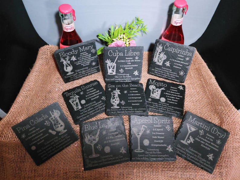 May include: A collection of dark gray slate coasters with engraved cocktail recipes, including "Bloody Mary," "Cuba Libre," and "Mojito." The coasters are arranged on a brown burlap surface, with two bottles of pink soda in the background.