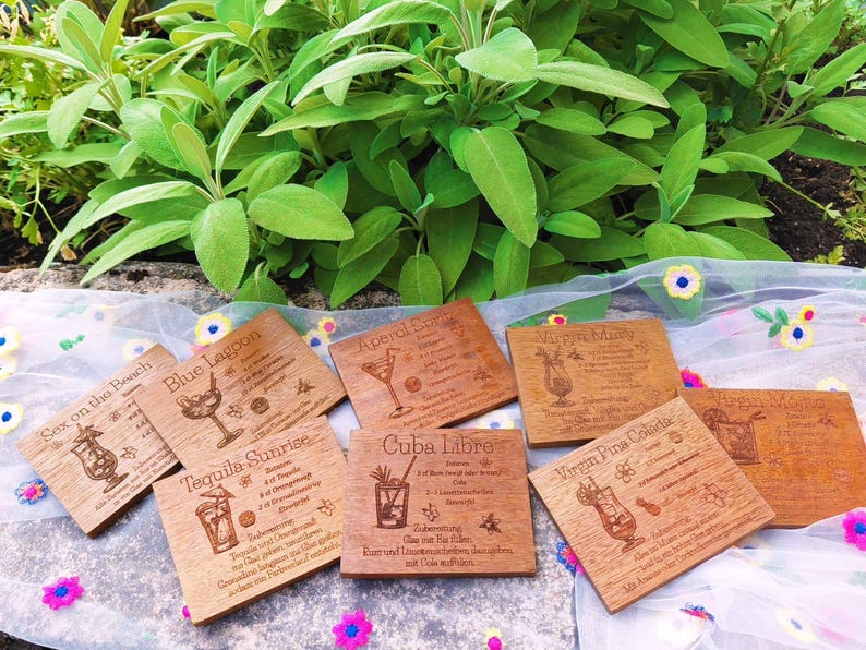 May include: Wooden coasters with engraved cocktail recipes, including "Sex on the Beach," "Blue Lagoon," and "Tequila Sunrise." The coasters are square, with a light brown finish, and are arranged on a floral patterned cloth with green foliage in the background.