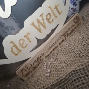 May include: Wooden sign with the words "der Welt" and "Mama" engraved. The sign also includes the phrase "im n&auml;chsten Leben w&uuml;nsche ich mir, dass du wieder meine Mutter wirst." in German. The sign is made of light-colored wood.