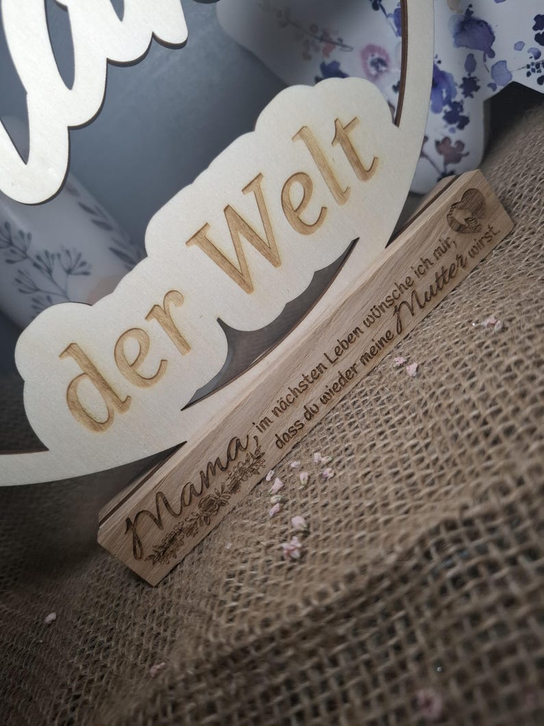 May include: Wooden sign with the words "der Welt" and "Mama" engraved. The sign also includes the phrase "im n&auml;chsten Leben w&uuml;nsche ich mir, dass du wieder meine Mutter wirst." in German. The sign is made of light-colored wood.